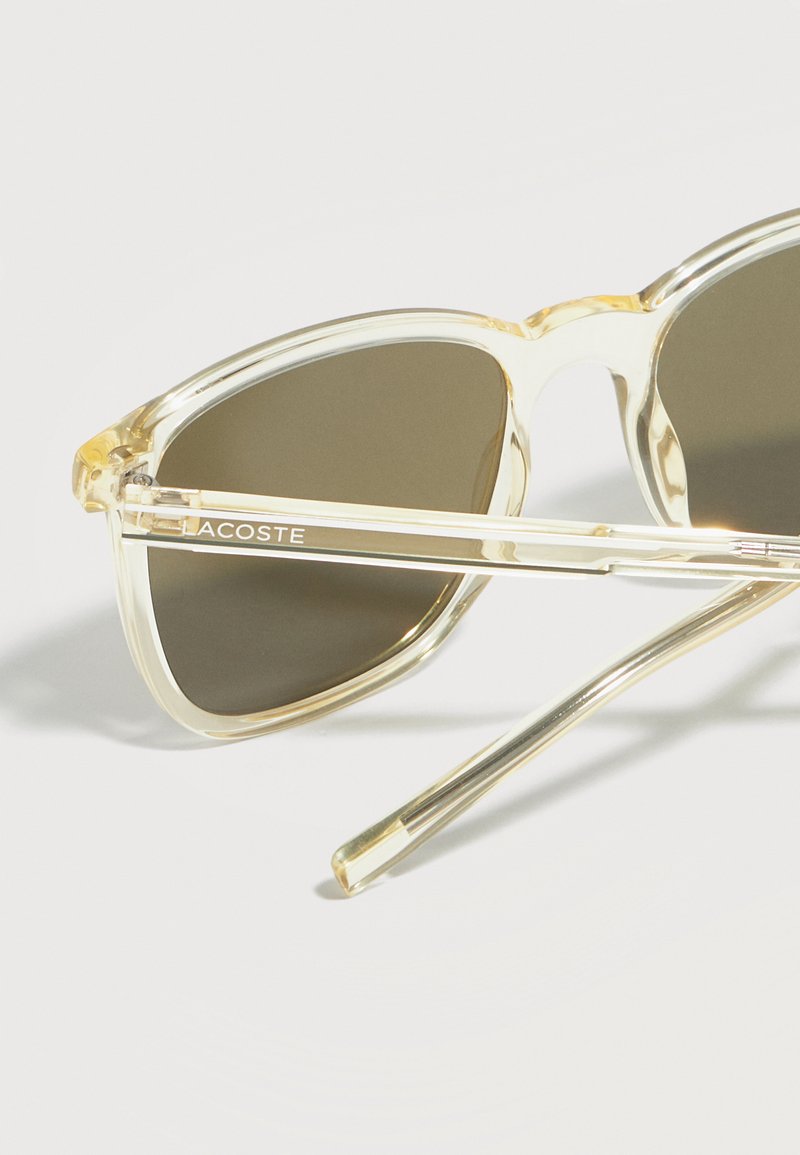 Occhiali da sole con montatura trasparente, lenti scure e il marchio "LACOSTE" sull'asta laterale, posizionati su una superficie bianca.