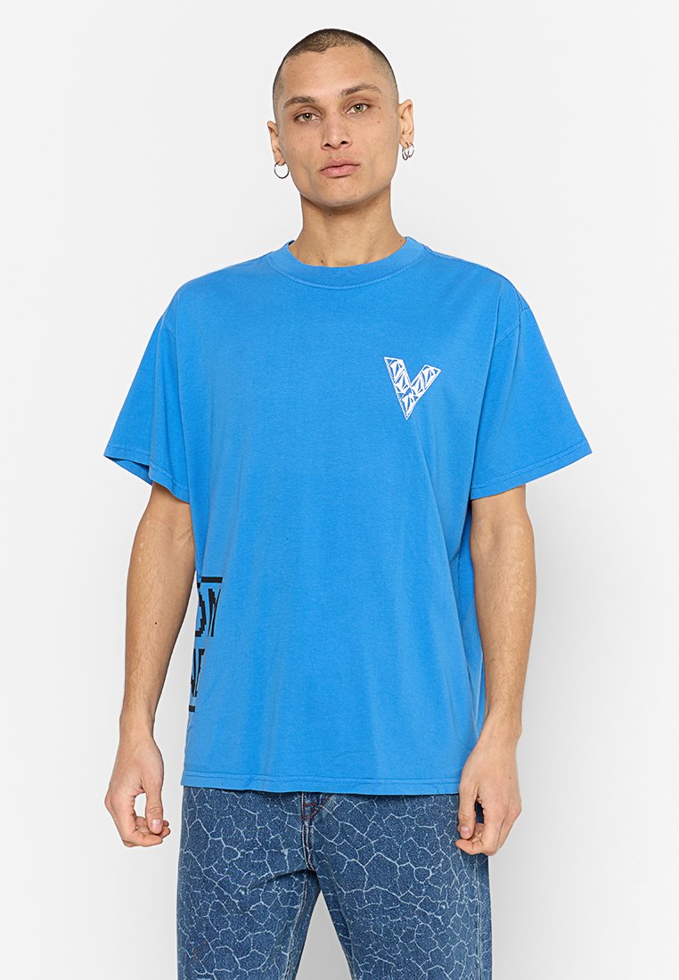 T-shirt en coton oversize bleu à manches courtes, avec un logo géométrique "V" blanc sur la poitrine et un texte graphique noir sur le côté.
