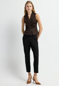 Brun ærmeløs bluse med en tætsiddende pasform og knapper nedad foran, parret med sorte tapered bukser og beige hæle.