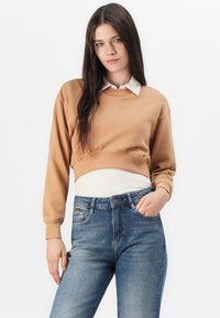 Avkortad sweatshirt i ljust brunt material med ribbat krage över en vit långärmad tröja, tillsammans med slim fit mörkblå jeans.