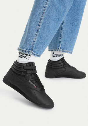 Person iført blå jeans, sorte Reebok høje sneakers med velcrolukninger og hvide Reebok logo sokker mod en ensfarvet hvid baggrund.