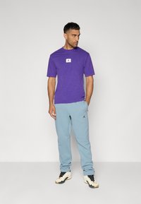 Lilla bomulds T-shirt med et hvidt logo-patch, parret med lyseblå sweatpants med et sort logo. Modellen har sorte og hvide sneakers på.