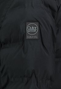 Schwarze Pufferjacke mit glatter Oberfläche, ausgestattet mit einem rechteckigen Logo-Patch, auf dem "Outerwear SUBLEVEL" in Grau und Weiß steht.