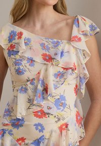 Γυναίκα που φοράει ένα αμάνικο κρεμ φόρεμα με μπλε, κόκκινο και κίτρινο floral μοτίβο, με στρωμένες πιέτες στο λαιμό και το μπούστο.