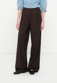 Brune vide benbukser med frontfolder og høj talje, lavet af et glat stof. Parret med en lyseblå button-up skjorte.