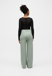 Moteris su garbanotais plaukais, vilkinti juodą nėriniuotą ilgomis rankovėmis viršutinį drabužį ir aukštu liemeniu plačiomis šviesiai pilkomis kelnėmis, stovinti priešais švarų foną, veidu atsigręžusi į šalį.