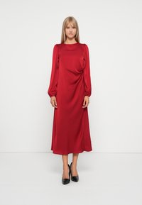 Robe midi en satin rouge avec de longues manches bouffantes et un col rond. Ourlet arrondi et texture lisse. Portée avec des chaussures à talons hauts noires.