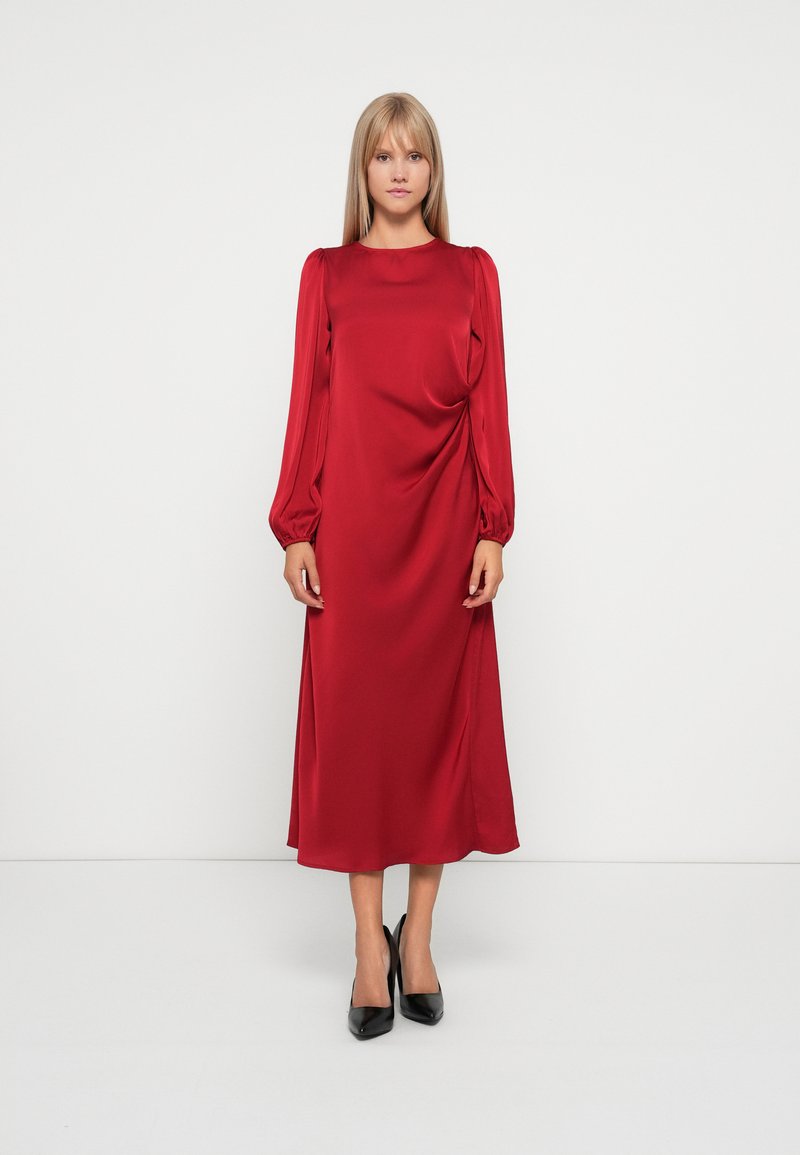 Robe midi en satin rouge avec de longues manches bouffantes et un col rond. Ourlet arrondi et texture lisse. Portée avec des chaussures à talons hauts noires.