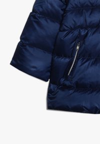 Granatowa pikowana kurtka z suwakiem na rękawie, na białym tle, ukazująca błyszczący, wypchany materiał oraz detale przeszycia.