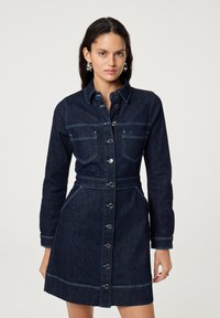 Vrouw met lang, donker haar draagt een donkerblauwe denim button-up jurk met voorzakken en zilveren ronde oorbellen, staand tegen een effen achtergrond.