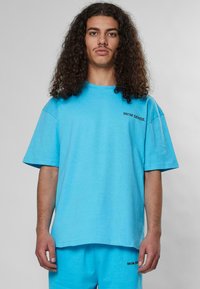 Trumpas rankovėmis, laisvas t-shirt ryškiai turkio spalvos medvilnės. Yra subtilus juodas logotipas kairėje pusėje. Lygi tekstūra ir neformalumas dizainas.