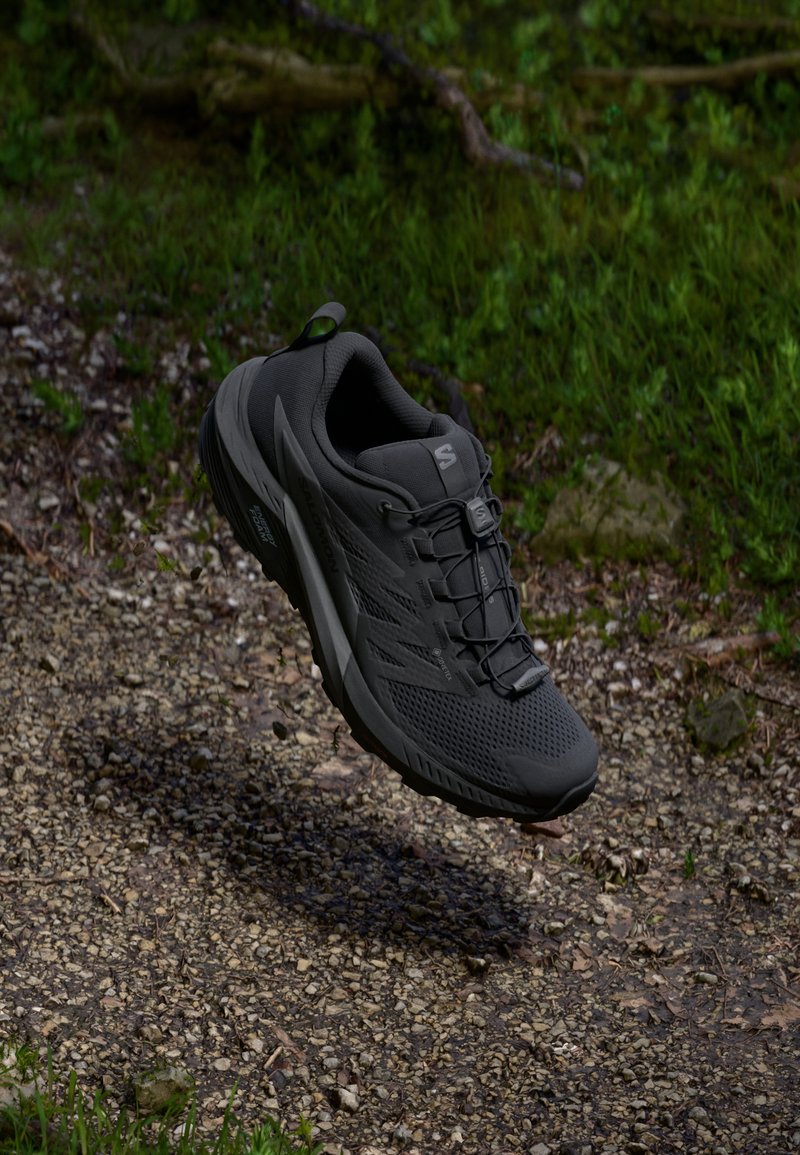Chaussure de sport noire avec une tige en maille texturée, un design léger et une semelle antidérapante, suspendue au-dessus d'un chemin de gravier.