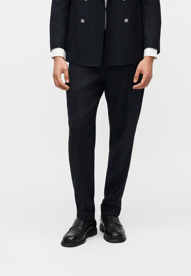 Άνδρας που φοράει μαύρο παντελόνι κοστουμιού ραμμένο στα μέτρα, μαύρα δερμάτινα μοκασίνια και μαύρο μονόπετο σακάκι με εμφανείς λευκές μανσέτες πουκάμισου.