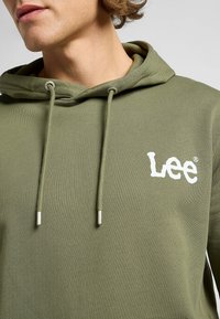 Felpa verde oliva con texture a coste, dotata di tasca frontale e lacci con accessori in metallo. Logo "Lee" bianco sul davanti.