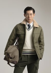 Chaqueta verde oliva con cuello beige, suéter blanco de punto trenzado y pantalones verde oscuro. Lleva un bolso marrón claro con detalles en marrón.