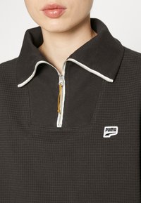 Gros plan d'une personne portant un pull à col montant Puma texturé de couleur sombre, avec une fermeture éclair blanche et un logo sur la poitrine.