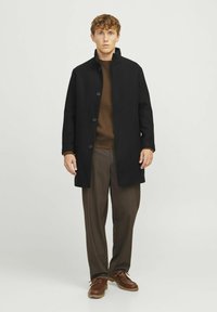 Abrigo negro hasta la rodilla con un alto collar y cierre de botones, usado sobre un suéter marrón y pantalones marrones. Zapatos marrones completan el atuendo.
