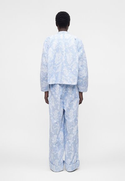 Άτομο με κοντά σγουρά μαλλιά φοράει ανοιχτό μπλε κεντημένο σακάκι και αντίστοιχο παντελόνι με φαρδιά πόδια, στέκεται ανάποδα μπροστά σε μονόχρωμο φόντο.