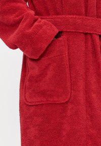 Punainen froteekylpytakki, jossa on teksturoitu pinta, vyötäröllä vyö ja vasemmalla puolella käytännöllinen tasku.