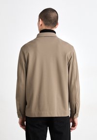 Beige, textured jakke med sort krave, knaplukning ved ærmerne og skræddersyet pasform, set fra ryggen mod en ensfarvet baggrund.