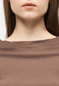 Εγγύς λήψη του κάτω μέρους του προσώπου και του λαιμού μιας γυναίκας που φοράει καφέ μπλούζα με φαρδύ λαιμό, με ίσια καστανά μαλλιά ορατά στις πλευρές.