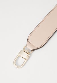 Vaalea beige nahkainen ranneke tummanruskeilla reunoilla, jossa on kullansävyinen metalliklipsi. Sileä pinta ja näkyvät kirjontalinjat.