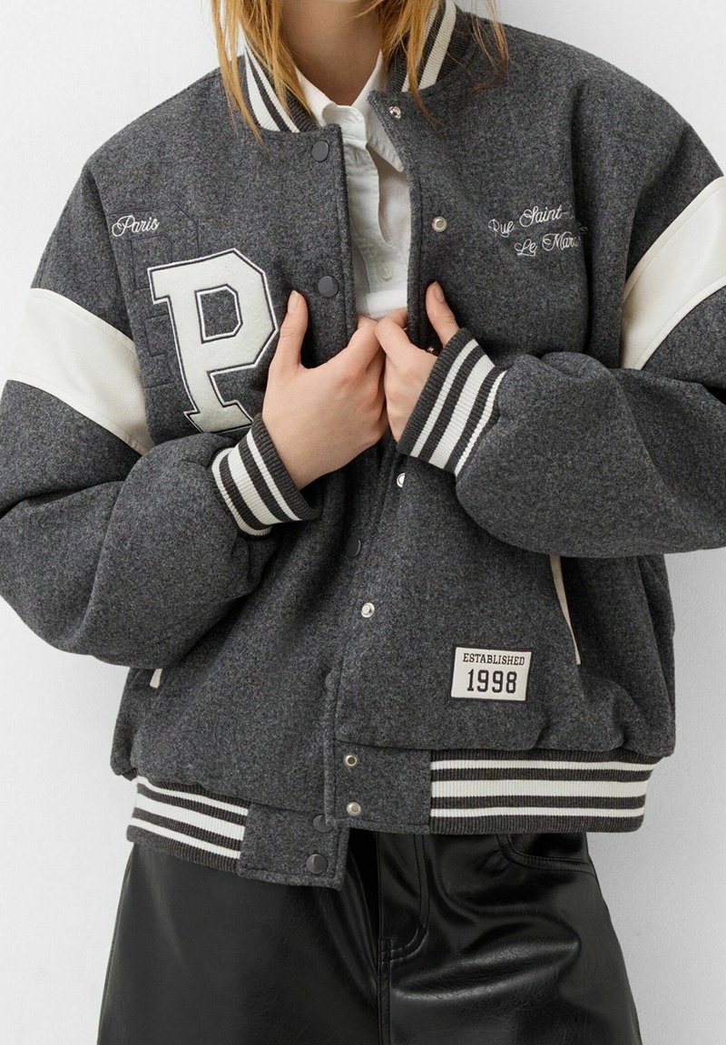 Personne attachant une veste varsity grise à rayures blanches, brodée des lettres "P", "Paris", "Établie en 1998", avec un col, portée sur une chemise blanche.