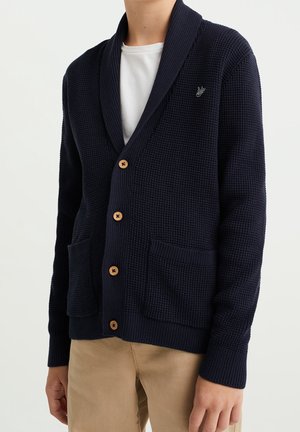 Mężczyzna noszący granatowy kardigan dziany w strukturę wafla z drewnianymi guzikami, dwie przednie kieszenie i białą koszulkę pod spodem, połączony z beżowymi spodniami.