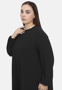 Sort langærmet kjole med rund halsudskæring. Stoffet er blødt med en glat overflade. Afsluttet med en kraftig perlekæde som accent.