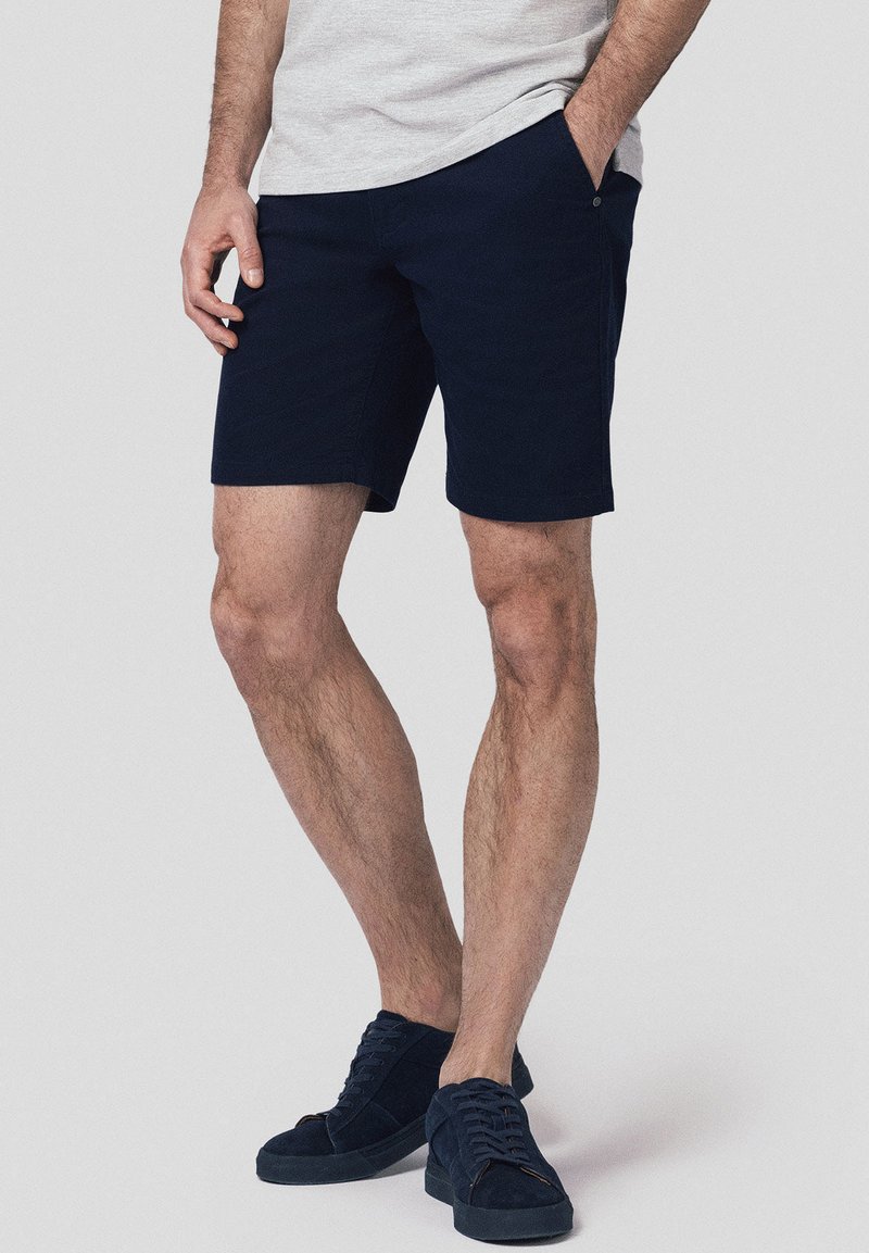 Granatowe bawełniane szorty o prostym kroju, z bocznymi kieszeniami i detalem w postaci guzika. Noszone z dopasowanymi granatowymi zamszowymi trampkami.