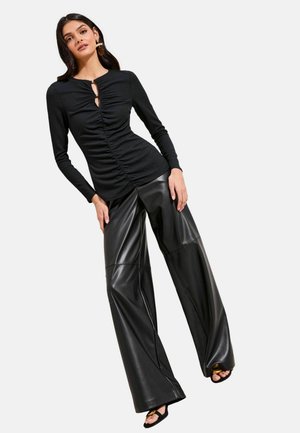 Žena oblečená v zvrásnenej čiernej blúzke s dlhými rukávmi a širokých nohaviciach z umelého koženého materiálu, pózujúca s jednou nohou mierne vpredu na svetlom pozadí.