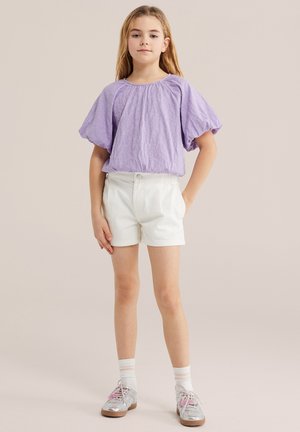 Mlada deklica stoji, oblečena v vijolično bluzo z napihnjenimi rokavi, bele kratke hlače, bele nogavice in srebrne čipkaste čevlje z roza poudarki.