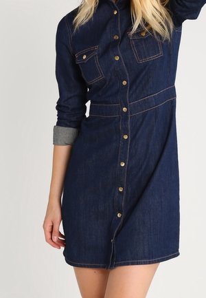 Kobieta ubrana w ciemnoniebieską sukienkę z denimu na guziki, z długimi rękawami, dwoma kieszeniami na piersiach i podwiniętymi mankietami.