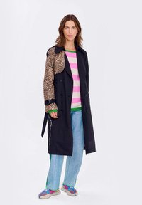 Čierny trenčkot s rukávom z leopardieho vzoru, s gombíkmi vpredu, prehodený cez ružovo-biele pruhované sveter, skombinovaný so svetlo modrými nohavicami a farebnými sandálmi.