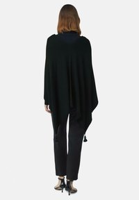 Donna in piedi di spalle, indossa un poncho asimmetrico nero con frange, pantaloni neri e sandali con tacco neri su sfondo bianco.
