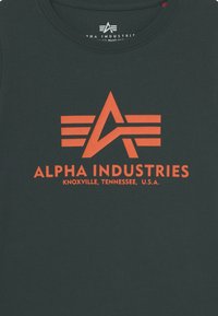 Tamsiai žalias medvilnės marškinėliai su oranžiniu grafiniu logotipu, kuriame pavaizduota žvaigždė ir horizontalios linijos, tekstas "ALPHA INDUSTRIES, KNOXVILLE, TENNESSEE, JAV."