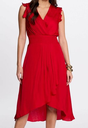 Rode mouwloze wikkeljurk met gerimpelde schouders, getailleerde taille en asymmetrische zoom, gedragen met dikke gouden armbanden aan de rechterpols.