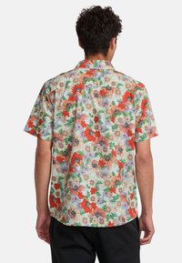 Short-sleeve button-up shirt featuring a vibrant floral pattern in red, orange, purple, and green on a light background, with a curved hem.