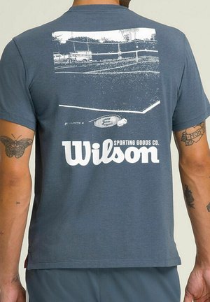 T-shirt bleu avec un motif blanc représentant un court de tennis, une raquette, une balle et le texte "Wilson Sporting Goods Co." au dos.