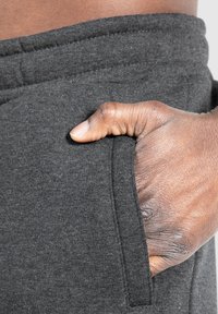Dunkelgraue Sweatpants mit einer weichen Textur, die eine Seitentasche und einen elastischen Bund aufweisen, teilweise gezeigt mit einer Hand, die in der Tasche ruht.