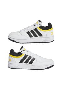Baskets basses blanches Adidas avec des bandes noires, des accents jaunes et des semelles noires, vues latérales orientées dans des directions opposées.