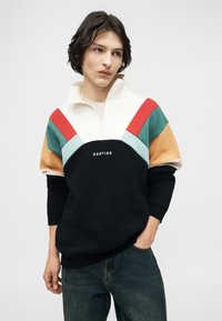 Νεαρός με κοντά σκούρα μαλλιά που φοράει πολύχρωμο φούτερ με φερμουάρ και την επιγραφή "KAOTIKO", ποζάροντας με το ένα χέρι στην τσέπη του τζιν.