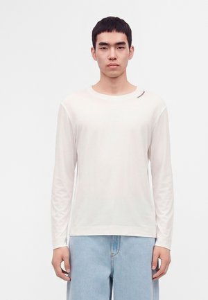 Ung mand med kort sort hår iført en almindelig hvid langærmet skjorte og lyseblå jeans, der står foran en hvid baggrund.