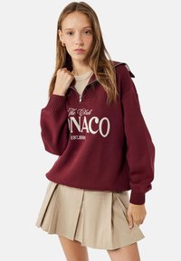 Sudadera burdeos con cremallera y puños y cuello acanalados, con "The Club Monaco EST.1991" impreso en blanco, combinada con una falda plisada beis.