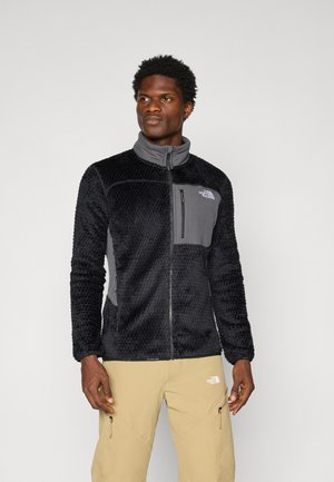 Black fleece jacket with textured fabric, gray panels, zippered chest pocket, and stand collar. The design features long sleeves.