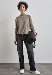 Maglione a maglia color marrone chiaro con collo alto e orlo arricciato; abbinato a jeans neri larghi e sneakers bianche; tiene in mano una borsa imbottita nera.