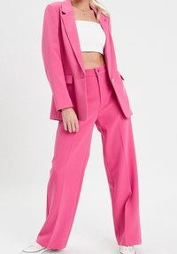 Kvinde iført en lys pink blazer og brede bukser med en hvid tube-top og hvide sneakers mod en ensfarvet baggrund.