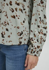 Blouse à manches longues en tissu vert clair, avec un imprimé animalier marron et doré. Poignets élastiqués, coupe décontractée et ourlet arrondi.