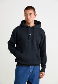 Sort hættetrøje lavet af bomuldsblanding, med snøre i hætten, en frontlomme og et kontrasterende hvidt Nike-logo på brystet.