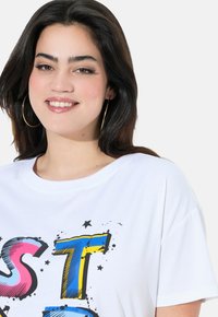 Camiseta blanca con un diseño gráfico colorido que presenta letras en negrita en rosa, azul y amarillo, acentuado con estrellas negras y efectos de salpicaduras.